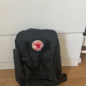 Svart ryggsäck från Fjällräven Kånken - Klassisk svart ryggsäck från Fjällräven Kånken med rund röd-vit logotyp framtill. Väskan har dragkedja, två handtag upptill och justerbara axelremmar. Väskan har en liten ficka där fram och sedan den stora med ett litet fack där ”sittunderlägget” brukar vara men som inte finns kvar. Väskan har även en stor ficka där bak med extra skydd som är menat för en dator eller iPad, en av de större modellerna. Väskan är i gott skick, pris går att diskutera och det är bara att höra av vid frågor🩷