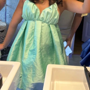 Mintgrön kortklänning med v-ringning - Säljer en mintgrön kortklänning med djup v-ringning och breda axelband. Klänningen har en luftig, utsvängd passform och är tillverkad i ett glansigt material som ger en lyxig känsla. Perfekt för sommarens tillfällen! 