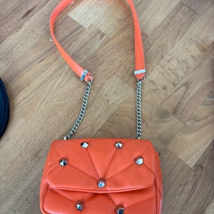 Orange axelväska med kedjedetalj - Säljer en orange axelväska med quiltad design och silverfärgade nitar. Väskan har en axelrem i syntet och en kedjedetalj i silver. Magnetlås och ett rymligt huvudfack. 