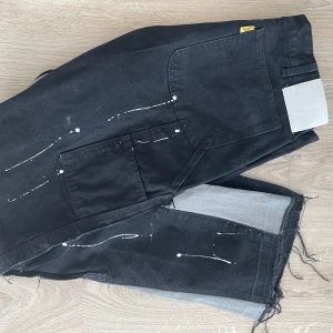 Svarta byxor med färgstänk från Gallery Dept. - Unika svarta byxor från Gallery Dept. med vita färgstänk och grå paneler längs benen. Byxorna har stora fickor på sidorna och råa kanter vid bensluten för en edgy look. Perfekta för dig som vill sticka ut med din stil.