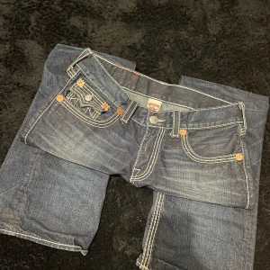 True religion jeans - Säljer ett par klassiska blå jeans från True Religion med tydliga vita kontrastsömmar och stora fickor med lock och knapp bak. Jeansen har normal passform och raka ben. Perfekta för dig som gillar streetwear och vill sticka ut lite extra.