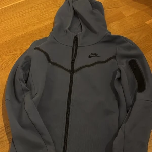 Blå Nike tech  - Säljer en blå  hoodiejacka  Nike tech  med dragkedja framtill och huva. Jackan har svarta detaljer och en ficka med dragkedja på ena ärmen. Perfekt för en sportig stil.