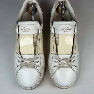 Valentino Garavani vita sneakers med beige detalj - Snygga vita sneakers från Valentino Garavani med en bred beige panel på sidan och klassisk snörning. Skorna har rund tå, platt sula och ikoniska nitar baktill. Perfekta för dig som gillar stilrena och exklusiva sneakers.