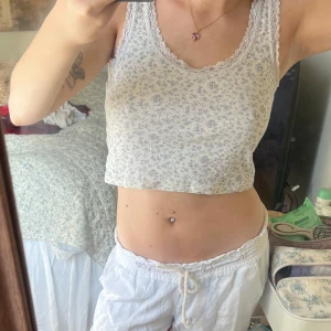 Vit croppad linnetopp med blommigt mönster - Söt vit croppad linnetopp från Brandy Melville i st S med små blommor i ljusgrått och spetskant runt halsringningen. Toppen har en tight passform och är ärmlös, perfekt för varma dagar.