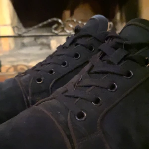 Christian Louboutins - Säljer ett par svarta sneakers från Christian Louboutin med klassisk röd sula. Skorna har snörning och är tillverkade i mocka med diskret broderad logga på hälen. Perfekta för dig som vill ha något stilrent men ändå sticker ut. Skorna är lite slitna men inga allvarliga skador 