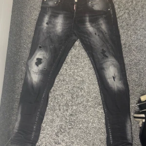 Svarta jeansbyxor från Dsquared2 ICON - Säljer ett par svarta Dsquared2 ICON jeans med slitna detaljer och hål. Byxorna har fem fickor, knappgylf och en smal passform. Snyggt tvättade partier och tydlig Dsquared2-logga bak på linningen.
