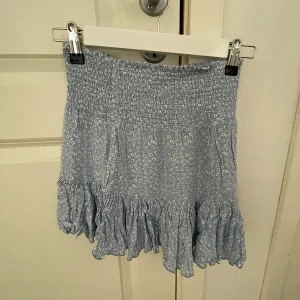 Ljusblå volangkjol med småblommigt mönster - Supersöt ljusblå kjol med små vita blommor och bred resår i midjan. Kjolen har volang nertill som ger en extra touch och är perfekt för varma dagar. Lätt och luftig modell som är enkel att matcha med en enkel topp. Snygg kjol skriv för fler bilder