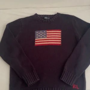 Mörkblå stickad tröja med amerikansk flagga från Ralph Lauren - Säljer en mörkblå stickad tröja från Ralph Lauren med en stor amerikansk flagga på bröstet och RL-broderi nertill. Tröjan har rund halsringning och långa ärmar med ribbade muddar. Storlek XL men den har krympt till S för att jag haft den i tvätten på hög värme 😁