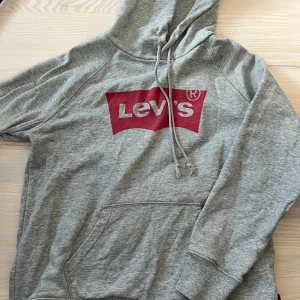 Grå hoodie från Levi's - Säljer en klassisk grå hoodie från Levi's med röd logga framtill. Tröjan har huva med dragsnöre och en stor magficka. Perfekt för en avslappnad och stilren look.