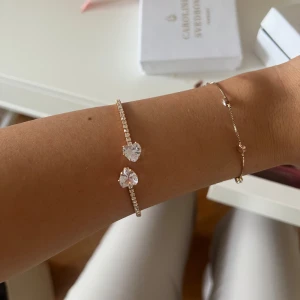 Guldfärgat armband med hjärtformade stenar - Säljer ett elegant guldfärgat armband med två stora hjärtformade, glittriga stenar och små gnistrande stenar längs bandet. Perfekt för att ge en lyxig touch till din outfit. Plastigt material som går att strecka ut långt om man vill att de ska sitta längre upp på armen 
