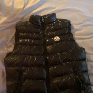  Moncler Väst -  Säljer en svart, glansig dunväst från Moncler med hög krage och dragkedja framtill. Västen har Moncler-logga på bröstet och en innerficka med röd dragkedja. Perfekt för lager-på-lager under kyliga dagar. Priset kan diskuteras 