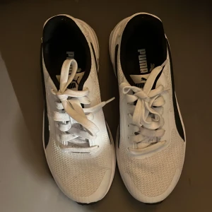 Vita sneakers från Puma med svarta detaljer - Säljer ett par fräscha vita sneakers från Puma med svarta detaljer på sidorna och sulan. Skorna har snörning och en sportig design med rund tå och platt sula. Perfekta för dig som gillar stilrena och bekväma skor. Helt oanvända.