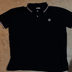 Mörkblå Moncler Pikétröja - Stilren och klassisk pikétröja från Moncler i mörkblått med ikoniska röda, vita och blå detaljer på krage och ärmslut. Märkesloggan är snyggt placerad på bröstet. Tröjan är i mycket fint skick och har endast använts ett fåtal gånger. Fick den i present men har inte riktigt kommit till användning 