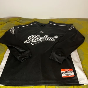 Harlem Oversized Sweatshirt - Säljer en svart långärmad tröja från Harlem med vitt tryck framtill och stort nummer 86 på ryggen. Tröjan har vita paneler på sidorna och detaljerade tryck på ärmarna. Perfekt för en sportig streetwear-look.