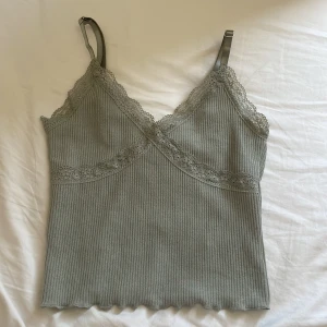 Grå grön ribbad linnetopp med spets från Divided - Supersöt grå ribbad linnetopp från Divided med spetsdetaljer längs urringning och axelband. Toppen har smala justerbara axelband och en croppad passform. Perfekt att styla till jeans eller kjol!