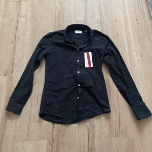 Svart skjorta från Lindbergh - Klassisk svart skjorta från Lindbergh med vit knappstängning och krage. Skjortan är långärmad och har en stilren, enkel design som passar till många tillfällen. Tillverkad i mjuk bomull för en bekväm känsla.