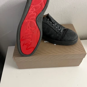 Gråa sneakers från Christian Louboutin - Säljer ett par gråa  sneakers från Christian Louboutin med klassisk röd sula. Skorna har snörning, rund tå och är tillverkade i mocka. Innersulan är beige och mjukt vadderad för extra komfort. Perfekta för dig som vill ha en stilren och lyxig look.