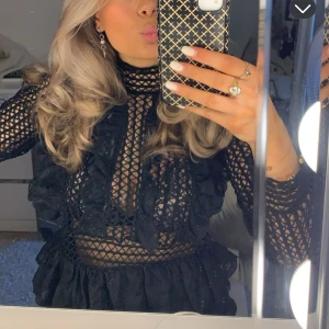 Svart spetstopp med volangdetaljer - Svart spetstopp med transparenta partier och spetsdetaljer. Volanger framtill och längs ärmarna. Hög hals och långärmad modell.