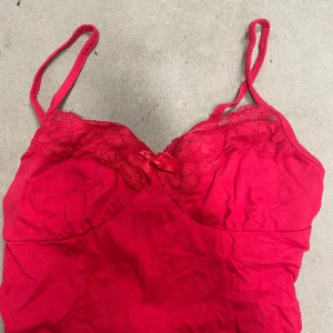 Röd spetsdetaljerad linnetopp från SHEIN - Säljer en röd linnetopp från SHEIN med tunna axelband, spets upptill och en liten rosett framtill. Toppen har en feminin och söt design, perfekt för att lyfta vilken outfit som helst.