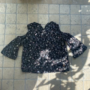 Svart blommig blus med öppna axlar - Säljer en svart blus med blommigt mönster i vitt, rosa och blått. Blusen har öppna axlar och vida trekvartsärmar. 