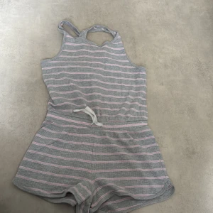 Grå och rosa randig kort jumpsuit - Superfin kort jumpsuit i grått med ljusrosa och vita ränder. Den har smala axelband som korsas i ryggen och ett knytband i midjan. Perfekt för varma dagar och riktigt skön att ha på sig.