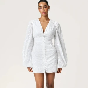 Vit kortklänning med v-ringning och puffärmar - Superfin vit kortklänning med djup v-ringning och långa puffärmar. Klänningen har knappar framtill och ett subtilt blommigt mönster i tyget. Perfekt för sommarens alla tillfällen! Storleken är 42 men har sytt in den i midjan, ärmarna och längden hos skräddade så den sitter ungefär som en 38!❤️