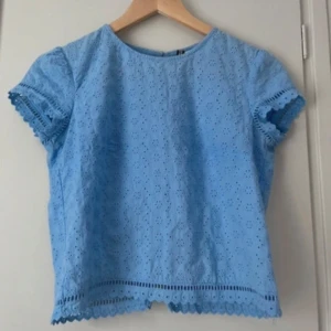 Ljusblå blus med spetsdetaljer - Söt ljusblå blus med korta ärmar och broderade spetsdetaljer. Blusen har rund halsringning och knappar längs ryggen. Perfekt för vår och sommar!