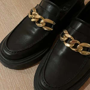Loafers i skinn med guldkedja 38 - Skinnskor från Duffy. Nyskick! Grov sula. Sköna på foten! Kraftig guldkedja. Stl 38.