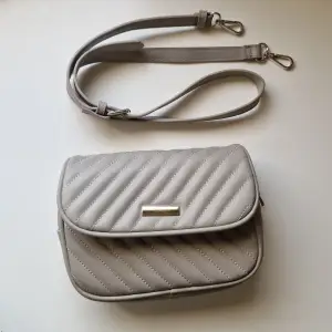 En grå handväska med dragkedja och magnetknäppe för att stänga den. Den är bekväm och enkel att ha med sig. Den har band man kan shystera längden på och man kan knäppa bort banden om man känner för det. Ser ut som ny och är inte sliten alls💕