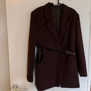 Kavaj med detaljer  - Oanvänd! Mörkbrun längre kavaj med detaljer. Den har en cut out i ena sidan, lite vaddering i axlarna och en mindre väska som går tvärs över kavajen. Priset är förhandlingsbart!