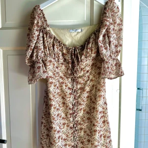 Blommig miniklänning - Jättegullig blommig klänning från NAKD!! ✨❤️‍🔥 Endast använt en gång så den är så gott som ny. Den är typ beige i undertonen med blommig mönster i gammelröd färg, ärmarna är lite puffiga och den har knytningsdetaljer längs hela framsidan. 