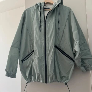 Grå vindjacka från Zara - Säljer en grå vindjacka från Zara i storlek M. Jackan har en dragkedja framtill, två fickor med dragkedja och en justerbar huva. Den är perfekt för vår och höst, och har en loose passform. Materialet är syntetiskt och jackan är i nyskick. Perfekt för att hålla vinden borta under kyliga dagar!