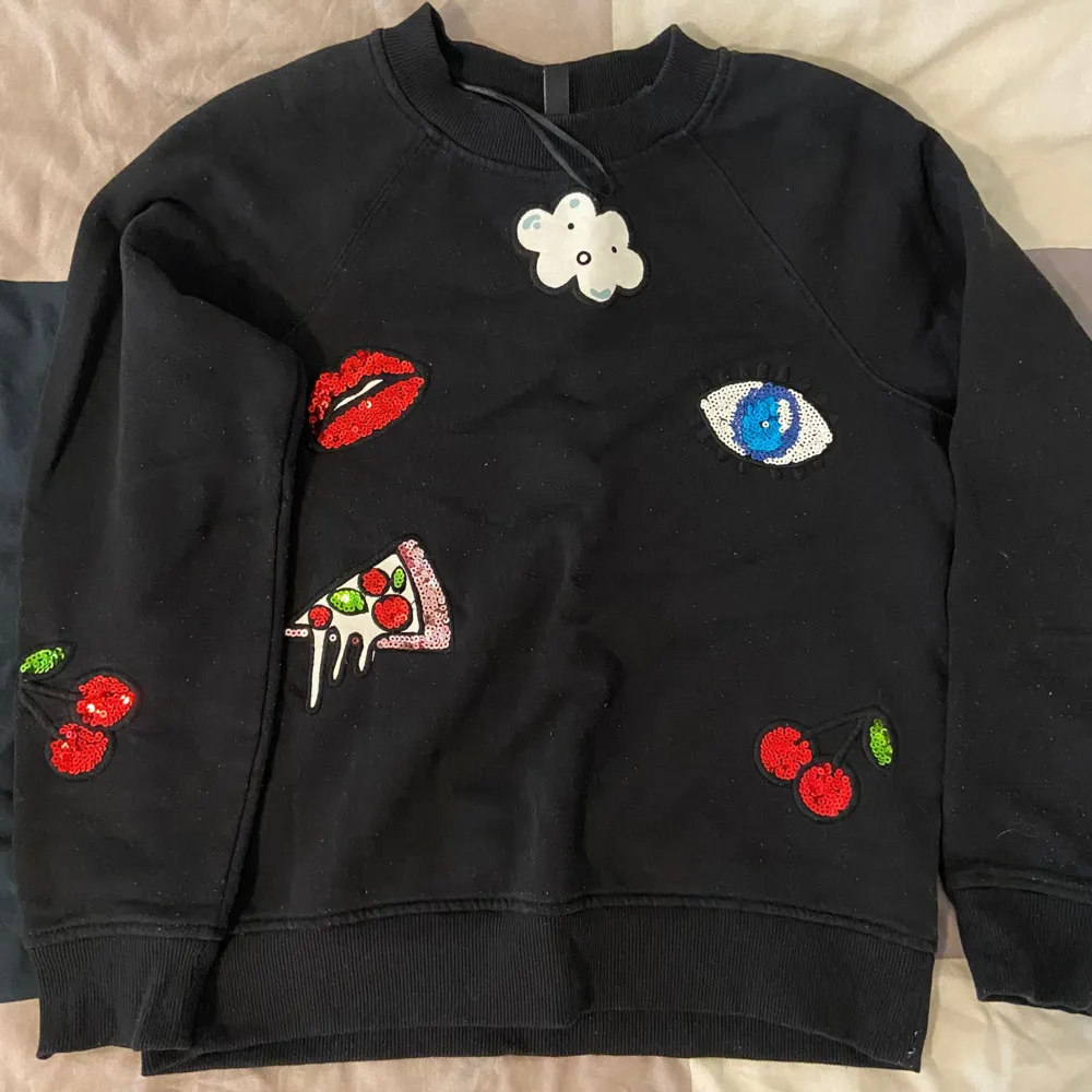 Säljer en svart sweatshirt från H&M Divided i storlek S. Tröjan har coola broderade detaljer som ett par röda läppar, en pizza slice, ett blått öga, körsbär och en söt liten molnfigur. Perfekt för en avslappnad och trendig look! 🖤🍒👁️🍕. Hoodies.