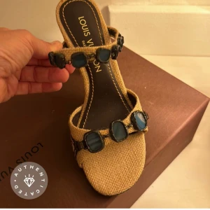 Beige klackskor från Louis Vuitton - Säljer ett par snygga beige klackskor från Louis Vuitton. Skorna har en öppen tå och är dekorerade med blå stenar på remmarna. De är tillverkade i ett textilmaterial som ser ut som jute och har en bekväm passform. Perfekta för sommarens alla fester!