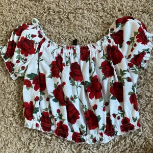 Blommig off-shoulder topp från H&M Divided - Säljer en superfin off-shoulder topp från H&M Divided. Den är vit med ett vackert rött blommönster och har korta ärmar. Toppen är croppad och har en elastisk kant vid axlarna och midjan för en bekväm passform. Perfekt för sommaren och festliga tillfällen! 🌸