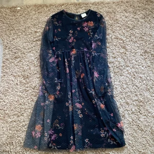 Blommig klänning från Zara Girls - Säljer en superfin blommig klänning från Zara Girls i storlek 10 (140 cm). Klänningen är mörkblå med ett vackert blommönster i olika färger. Den har långa ärmar och en söt liten rosett på ärmslutet. Perfekt för festliga tillfällen eller vardagsbruk. Klänningen är i nyskick och har en normal passform. Materialet känns lätt och luftigt, vilket gör den bekväm att bära.