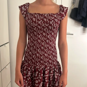 Vinröd blommig klänning - Säljer en superfin vinröd klänning med blommigt mönster. Klänningen är kort och har en söt volangdetalj längst ner. Den kan bäras både off-shoulder och med axelband. Perfekt för sommarens fester eller en mysig vårdag. Klänningen är i nyskick och passar perfekt för en festlig tillställning.💞❤️