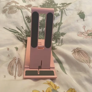 Rosa justerbart mobilställ - Säljer ett rosa justerbart mobilställ som är perfekt för att hålla din mobil i olika vinklar. Stället har svarta gummidetaljer för att hålla mobilen på plats och förhindra att den glider. Det är kompakt och lätt att ta med sig, perfekt för både hemmet och kontoret.
