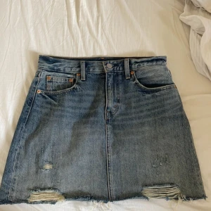 Jeanskjol från Levi's - Säljer en snygg jeanskjol från Levi's i storlek 27 (motsvarar dam S). Kjolen är i en klassisk blå jeansfärg och har en cool distressed look med slitningar och fransig nederkant. Perfekt för en avslappnad och trendig stil. Den har fem fickor och knappar fram. Passar perfekt till både vardag och fest!