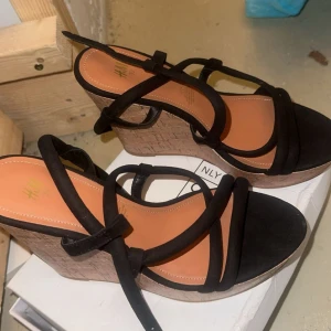 Svarta kilklackar från H&M - Säljer ett par snygga svarta kilklack från H&M i storlek 39. Skorna har en bekväm kilklack i kork och svarta remmar som ger ett stilrent och elegant utseende. Perfekta för vardag samt alla fester och tillfällen