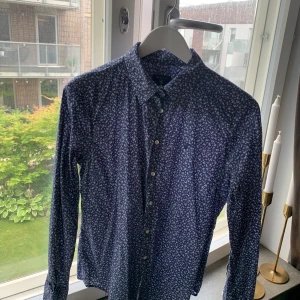 Blå blommig skjorta från Gant - En stilren blå skjorta från Gant med ett diskret blommönster i vitt. Skjortan har långa ärmar och knappar framtill. Perfekt för både vardag och fest.