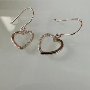 Hjärtformade öronhänge  - BESKRIVNING  Örhängen i äkta silver i form av hjärtan med små klara Cubic Zirconia stenar upptill. Ett par nätta örhängen med stilren design och skimrig detalj.
