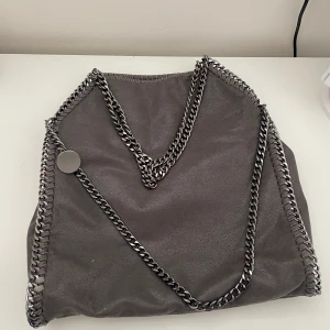 En grå väska med kedjor - Säljer en grå väska med kedjor, likt dem andra som är dyra. Den har aldrig kommit till användning och har därför bara varit i min garderob därav lite skrynklig men det går bort efter hand som man använder väskan🩷