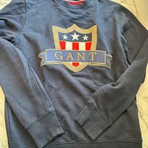 Mörkblå Gant Tröja  - En mörkblå Gant tröja i storlek S med ett stort gant tryck i mitten. Tröjan är av de mjukare materialen och är lätt att röra sig i. Passformen är ganska tajt runt ärmarna men lösare i mitten men perfekt runt midjan. Tröjan har endast använts 5-10 ggr
