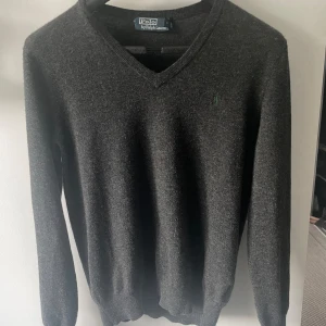 Grå tröja från Polo by Ralph Lauren - En stilren grå tröja från Polo by Ralph Lauren med v-ringning och långärmade ärmar. Tröjan har en broderad grön logotyp på bröstet och är gjord av 100% merinoull 