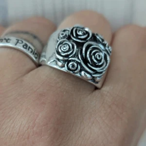 Silverfärgad ring med rosor - En vacker silverfärgad ring dekorerad med detaljerade rosor. Ringen har en bred design och är prydd med flera rosor i olika storlekar, vilket ger den ett unikt och elegant utseende.