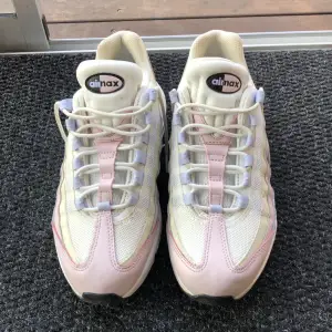 Ett par Nike Air Max sneakers i vitt, rosa och lila. Skorna har en klassisk Air Max-design med synlig luftdämpning i sulan och snörning framtill. De är tillverkade i en kombination av mesh och syntetmaterial för ökad komfort och andningsförmåga.