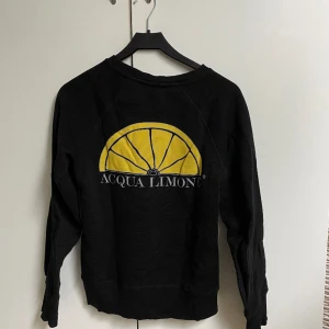 Svart sweathirt med tryck på ryggen - En svart sweathirt från Acqua Limone. Den har en gul citron på ryggen och är i nyskick. Den är i storlek XS och jag är 170cm lång.
