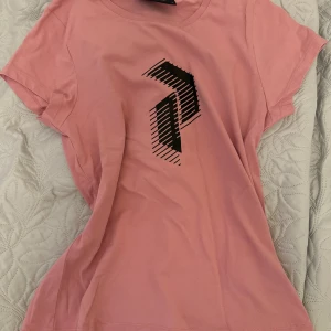 Rosa t-shirt från Peak Performance - En OANVÄND rosa t-shirt från Peak Performance med ett svart grafiskt tryck på framsidan. T-shirten har korta ärmar och är tillverkad i bomull.