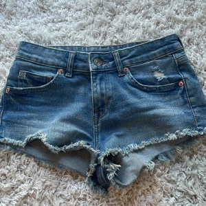 Blå jeansshorts från H&M Divided - Ett par blå jeansshorts från H&M Divided i storlek EUR 34. Shortsen har en lågmidjad passform och är tillverkade i slitstarkt denim med slitna detaljer och fransiga kanter. De har fem fickor och stängs med knapp och dragkedja.